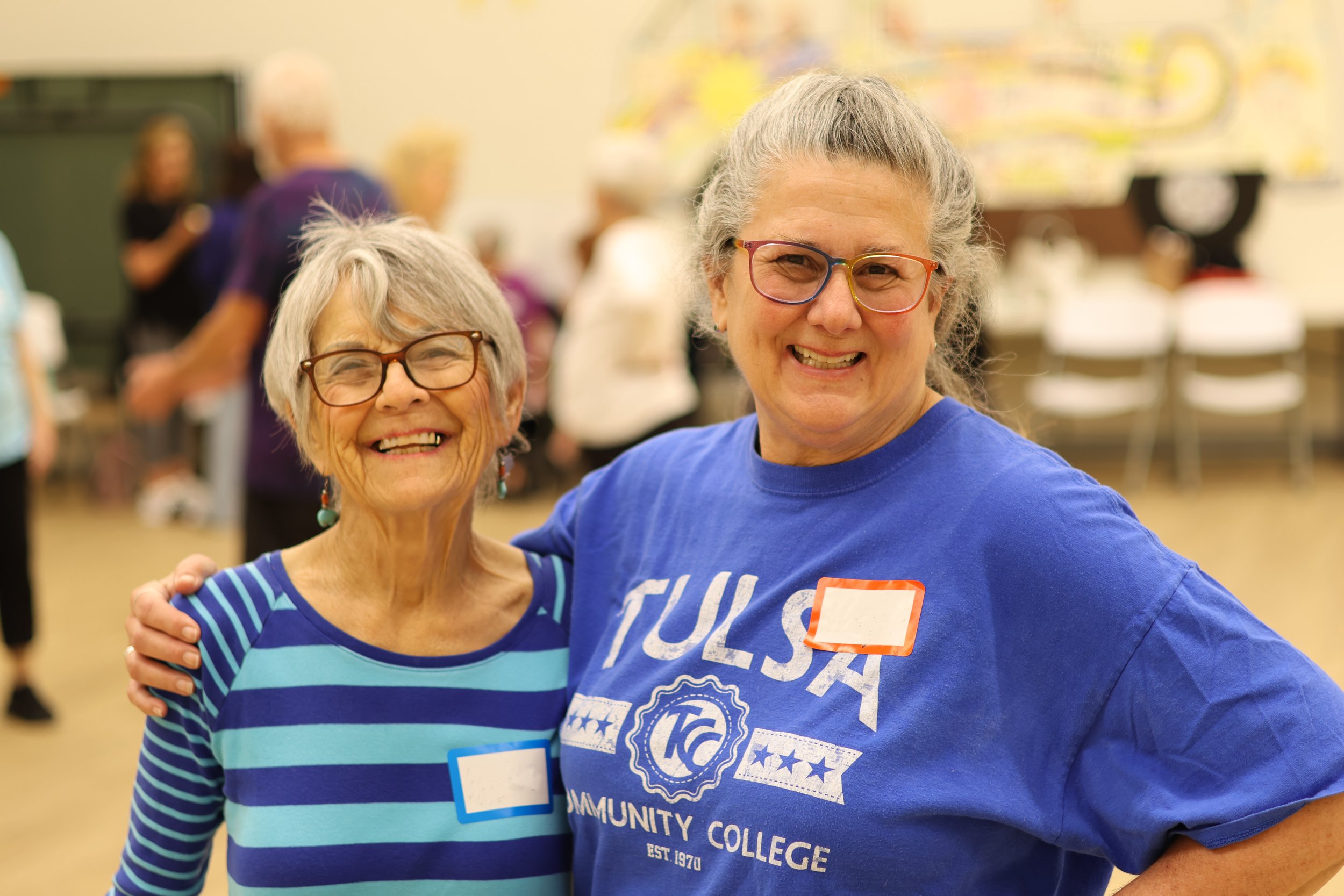 Two smiling women standing in a communal indoor setting with name tags, one wearing glasses and a blue striped top, the other in a blue "Tulsa Community College" T-shirt.