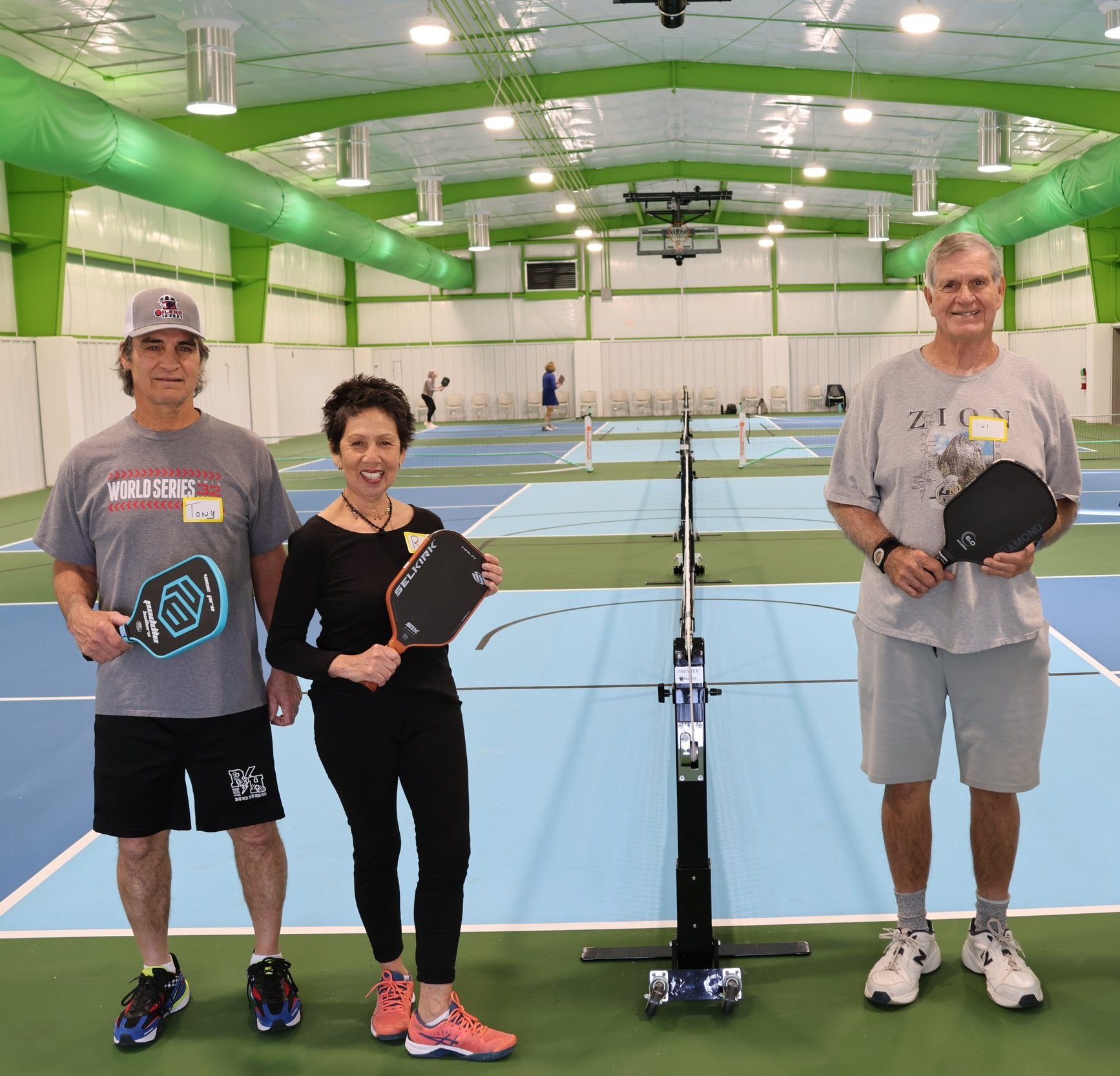 Three people holding pickleball paddles standing on an indoor court with multiple pickleball courts visible in the background.