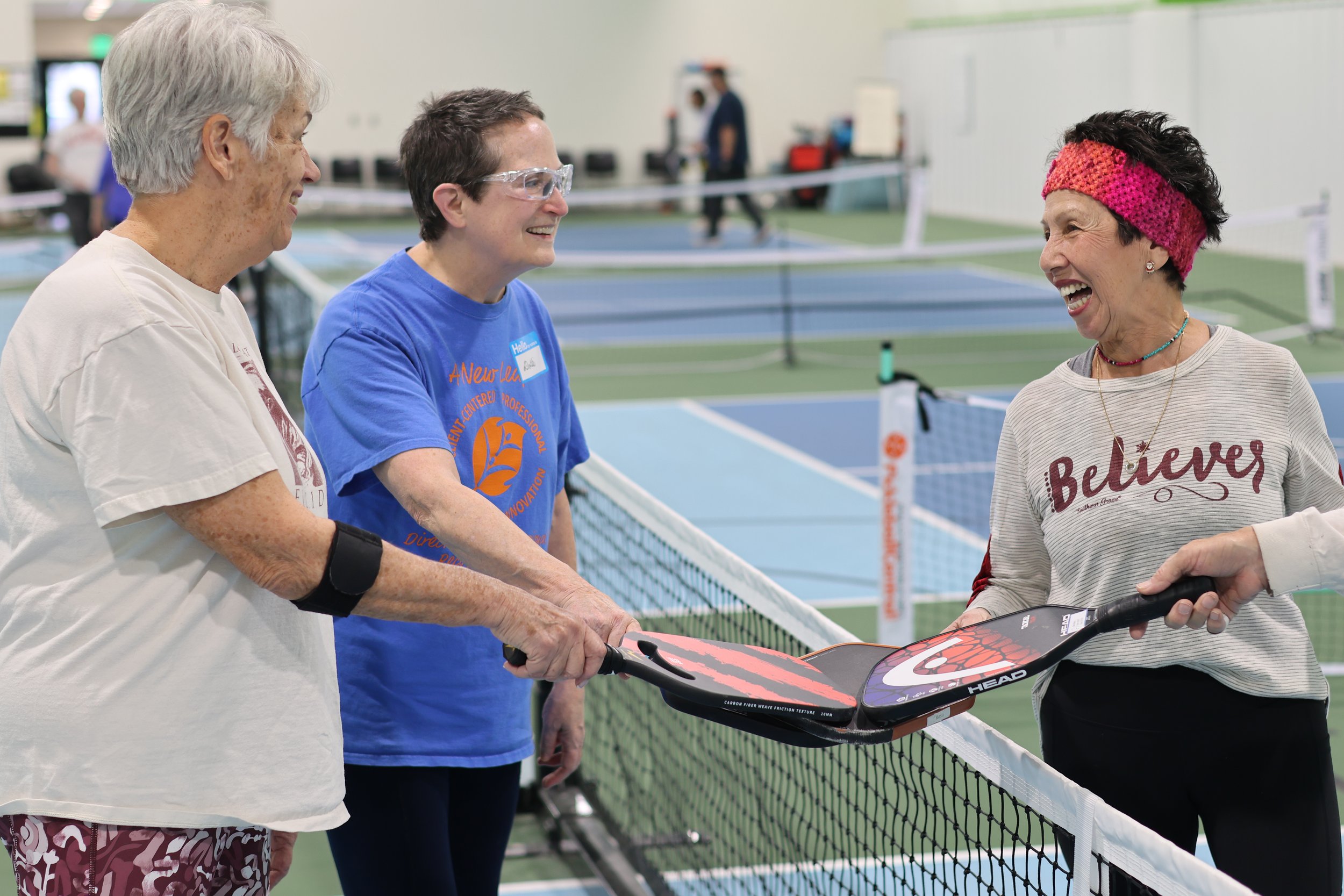 Three women with pickleball paddles smiling and talking on an indoor court.