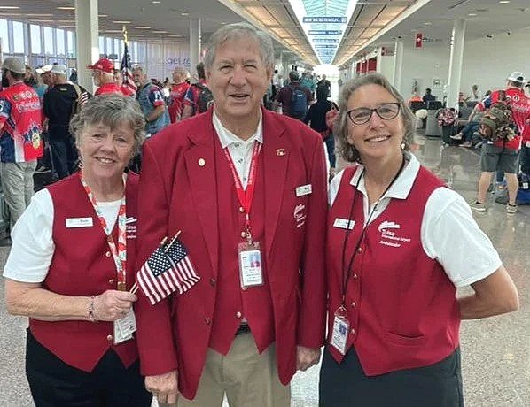 Three happy volunteers in red and white uniforms posing together at an airport, with a crowd in the background.