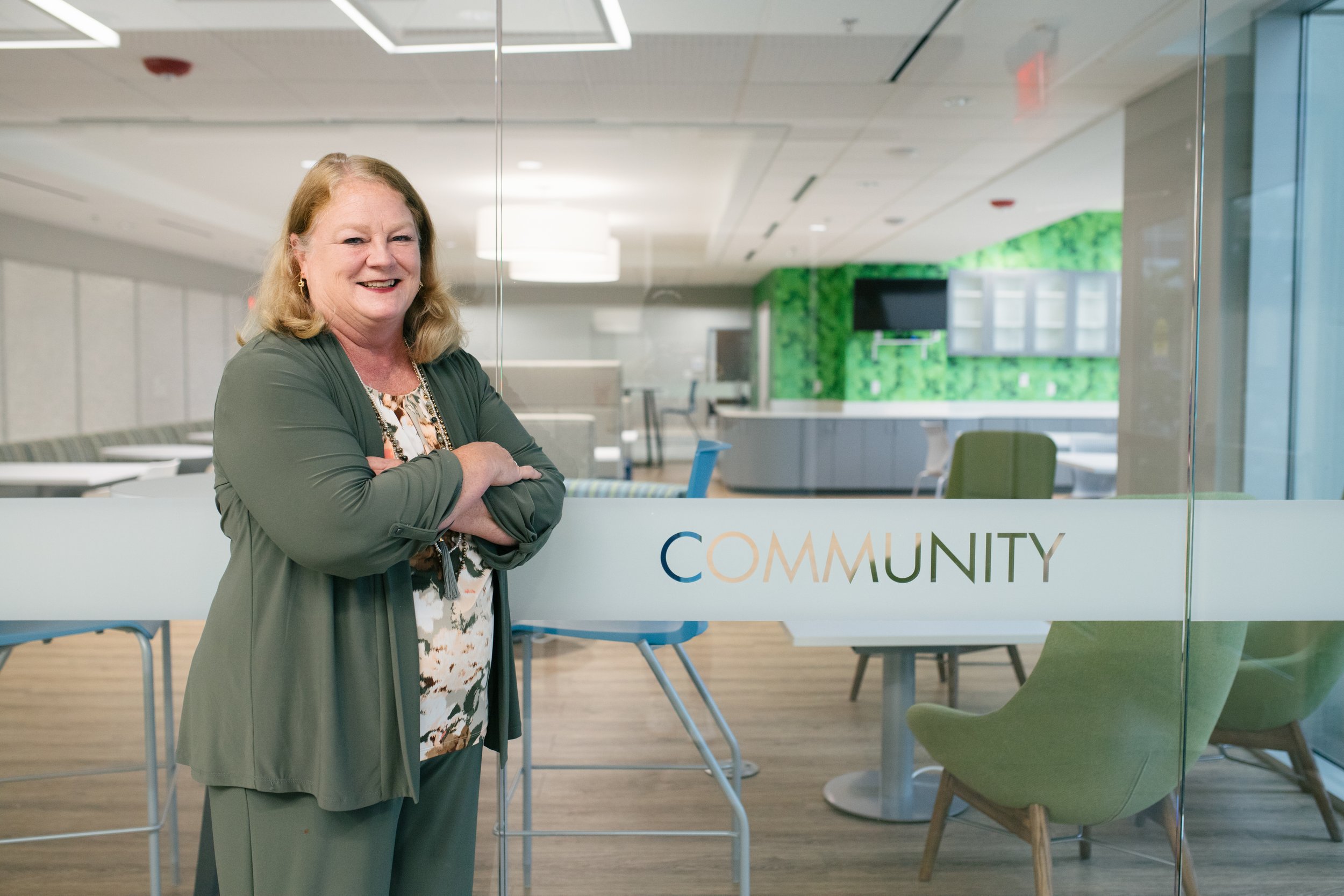 CEO Eileen Bradshaw LIFE Senior Services in a green blazer and patterned blouse standing with her arms crossed in front of a community room with green and white decor, chairs, and a glass wall with the word 'COMMUNITY' on it.