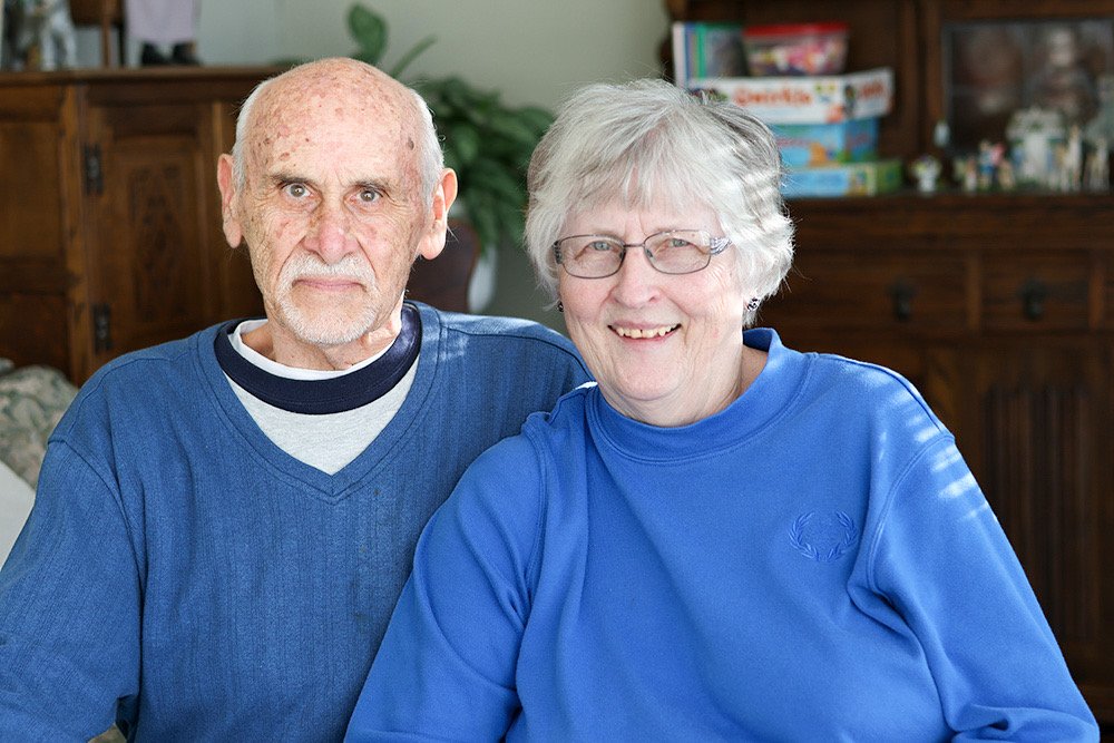 Elderly couple seated together in a room with board games in the background.
