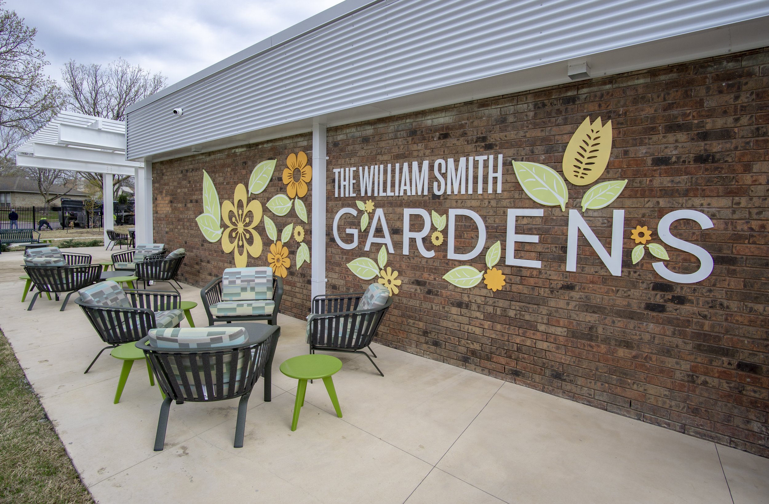 Outdoor seating area with chairs and tables, brick wall with decoration and text "The William Smith Gardens," featuring floral and leaf designs.