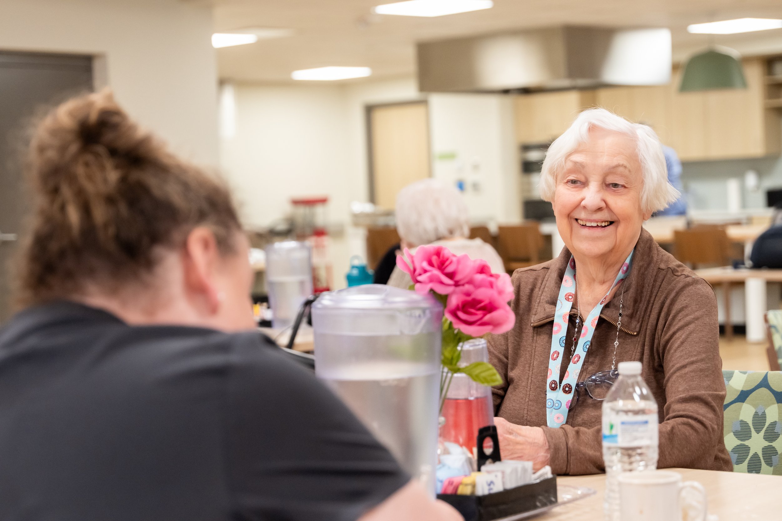 An elderly woman in Adult Day Health and a younger woman sitting at a table in a community center or cafeteria.