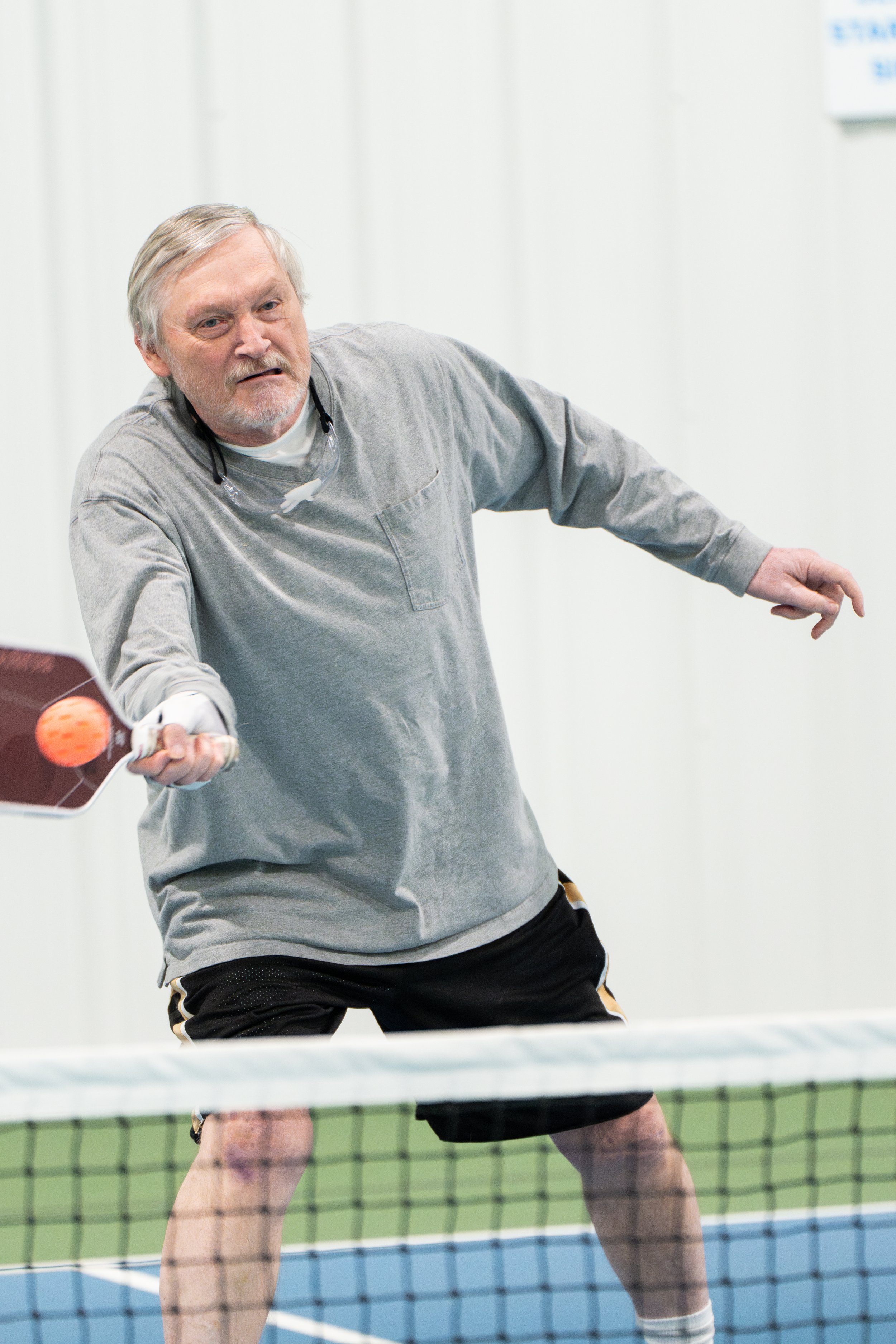 Elderly man playing pickleball indoors, hitting ball with paddle